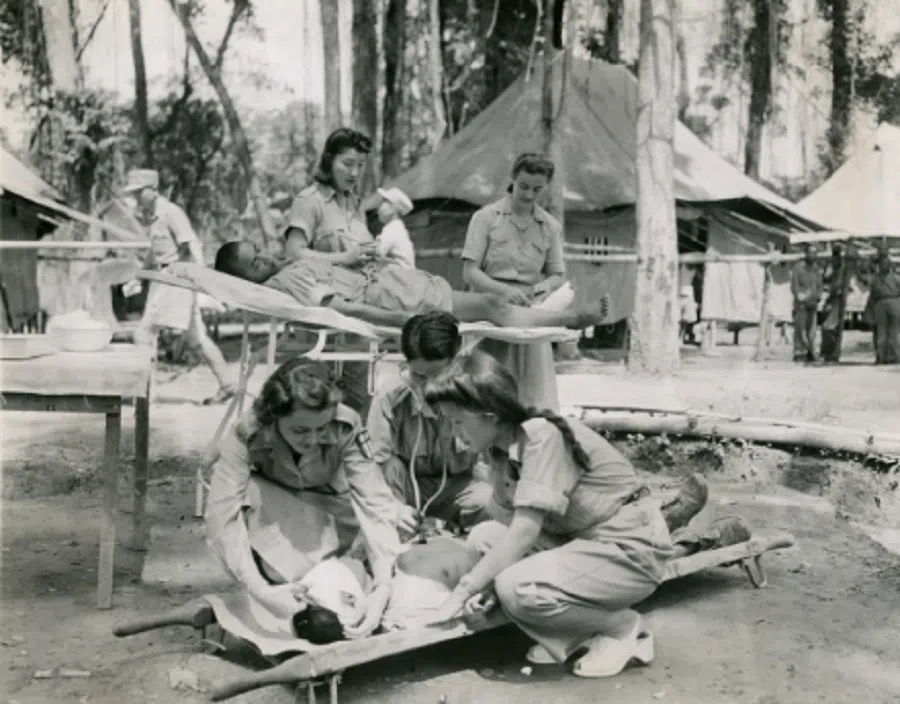 A large vintage photograph dated 14 October 1943 captures Nancy Cooke, a nurse from Philadelphia, Pennsylvania, dressing the head wound of an injured Chinese soldier at a field hospital in northern Burma. Behind her is her fellow nurse, Marie M. Carroll, also from Philadelphia. These wounded soldiers are from the battles in the Naga Hills, marking the first battle of the northern Burma counteroffensive and the first time that Chinese and American forces fought side by side.