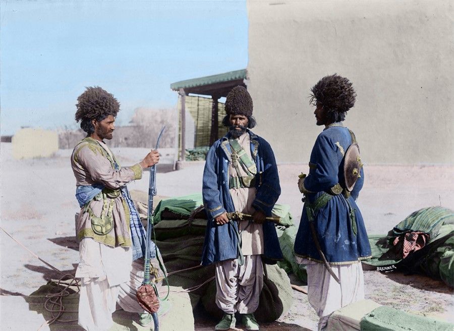 Afghanistan, 1890. Three Afghan men are wearing long pants and shirts that used to be white but are now sandblown and weathered. Over their shirts are blue jackets, and on their heads black sheepskin hats, the style favoured by Central Asian men. They are all clasping weapons for self-defence, as they stand guard over their wares.