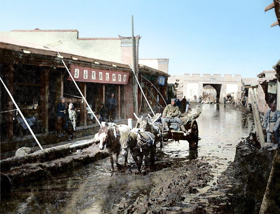 Urumqi, Xinjiang, 1930s. A man drives a horse cart on a muddy road in Nanmen (Akkowuk) district; some people are wearing light shirts, while others are wearing thick woollen jackets. The buildings on either side are flat-roofed, an architectural feature in desert regions. Urumqi was a way station on the way to Central Asia, where merchants and various groups on the Silk Road rested and exchanged information, a gathering point for various cultures.