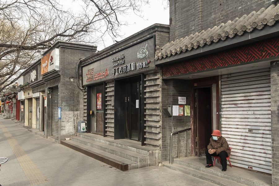 An elderly person wearing a protective masks sits in front of shuttered stores near a hutong neighborhood in Beijing, China, on 18 March 2020. (Qilai Shen/Bloomberg)