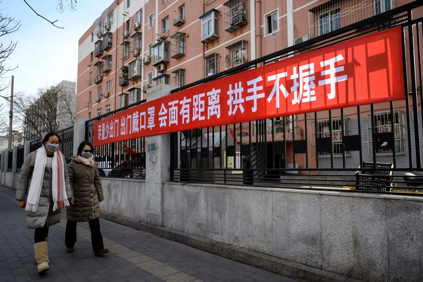 This photo taken on 1 February 2020 shows people wearing face masks walking at a compound in Beijing with a banner reminding people to reduce going outside, wear masks, and wash hands. (Photo by Noel Celis/AFP)