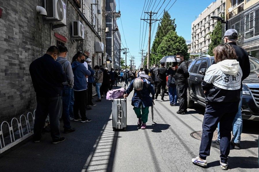 An elderly woman with luggage walks in an alley as people line up to be tested for Covid-19 outside a hospital in Beijing on 13 May 2022. (Jade Gao/AFP)
