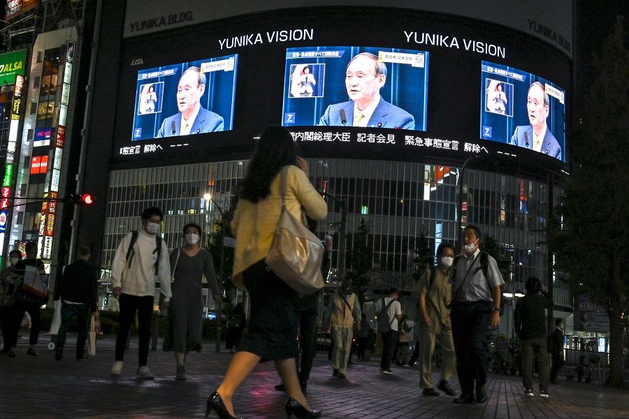 Pedestrians walk on a street and watch a live TV broadcast of then Japanese Prime Minister Yoshihide Suga announcing the lift of the coronavirus state of emergency in Tokyo's Shinjuku area on 28 September 2021. (Kazuhiro Nogi/AFP)