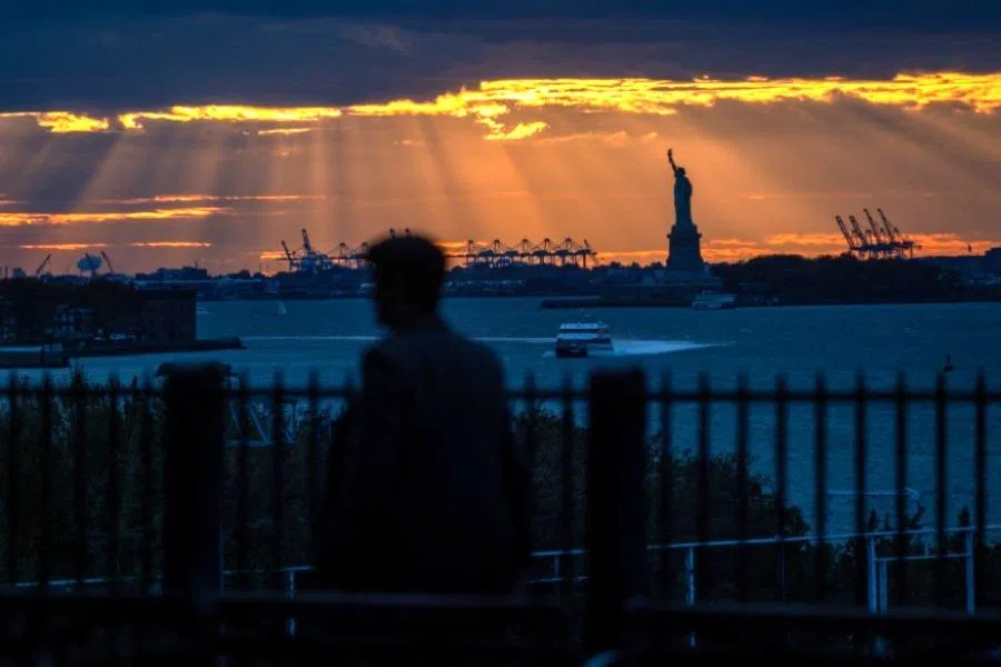 People stand on the Brooklyn promenade at sunset as the Statue of Liberty stands in New York Harbor on 22 October 2025 in New York City.