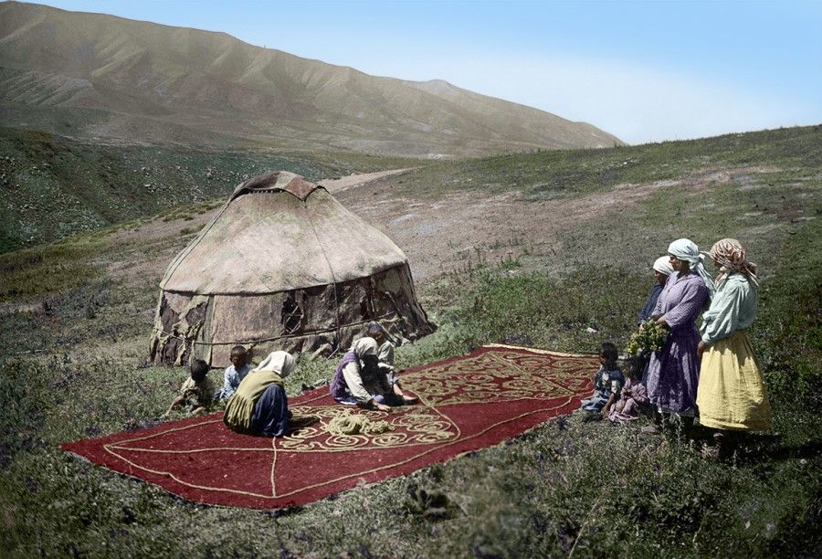 Uzbekistan, early 20th century. Two elderly women sit cross-legged on a carpet, preparing materials for weaving, as younger womenfolk stand to one side watching and learning about the weaving process from their elders.