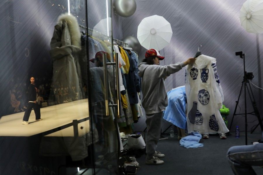 A salesperson promotes clothing by XUNRUO during a livestreaming session inside a booth while a model onstage presents creations from CHUSAN designed by Guoqin Zhang during China Fashion Week in Beijing, China, 31 March 2021. (Tingshu Wang/Reuters)
