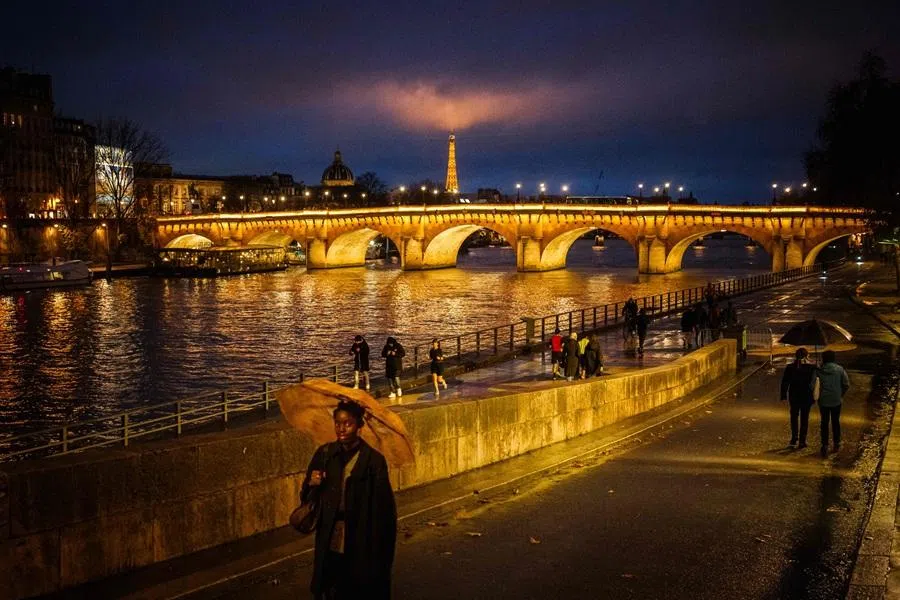 People walk along the banks of the Seine River with an illuminated Pont Neuf and the Eiffel Tower seen on the backgraund in Paris on 29 November 2025. (Dimitar Dilkoff/AFP)
