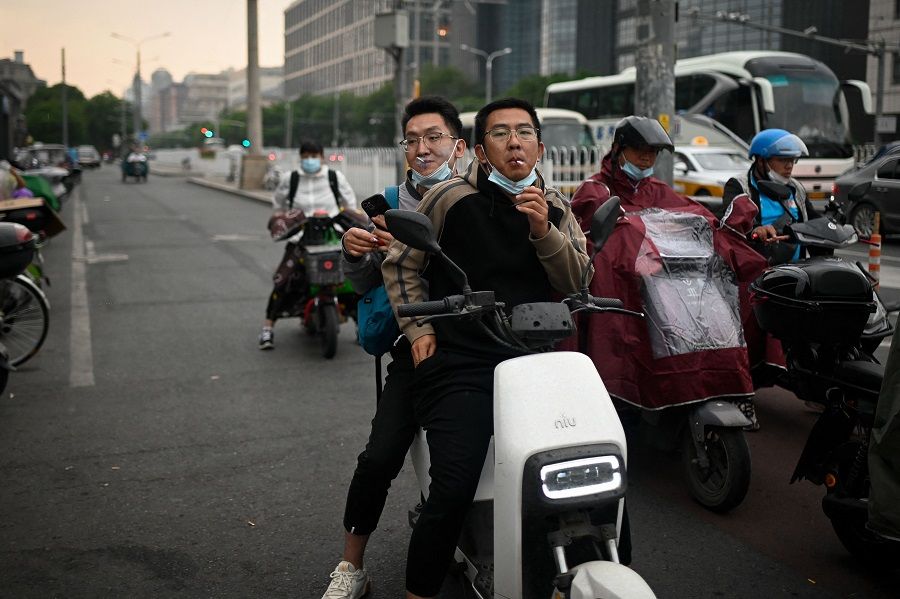 Two men riding an electric scooter smoke cigarette as they wait to cross a street in Beijing, China, on 12 May 2023. (Wang Zhao/AFP)