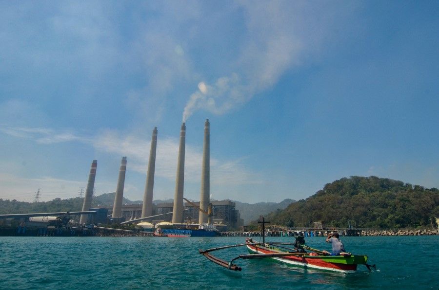 This photo taken on 22 September 2021 shows fishermen on their boat as smoke rises from chimneys at the Suralaya coal power plant in Cilegon. (Ronald Siagian/AFP)