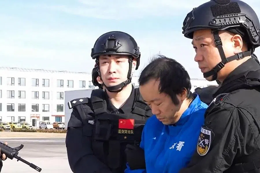 This frame grab taken from undated handout video footage released by China's Ministry of Public Security on 8 January 2026 shows guards escorting handcuffed accused scam boss Chen Zhi (C) off a China Southern plane in Beijing. (Handout/China’s Ministry of Public Security/AFP)