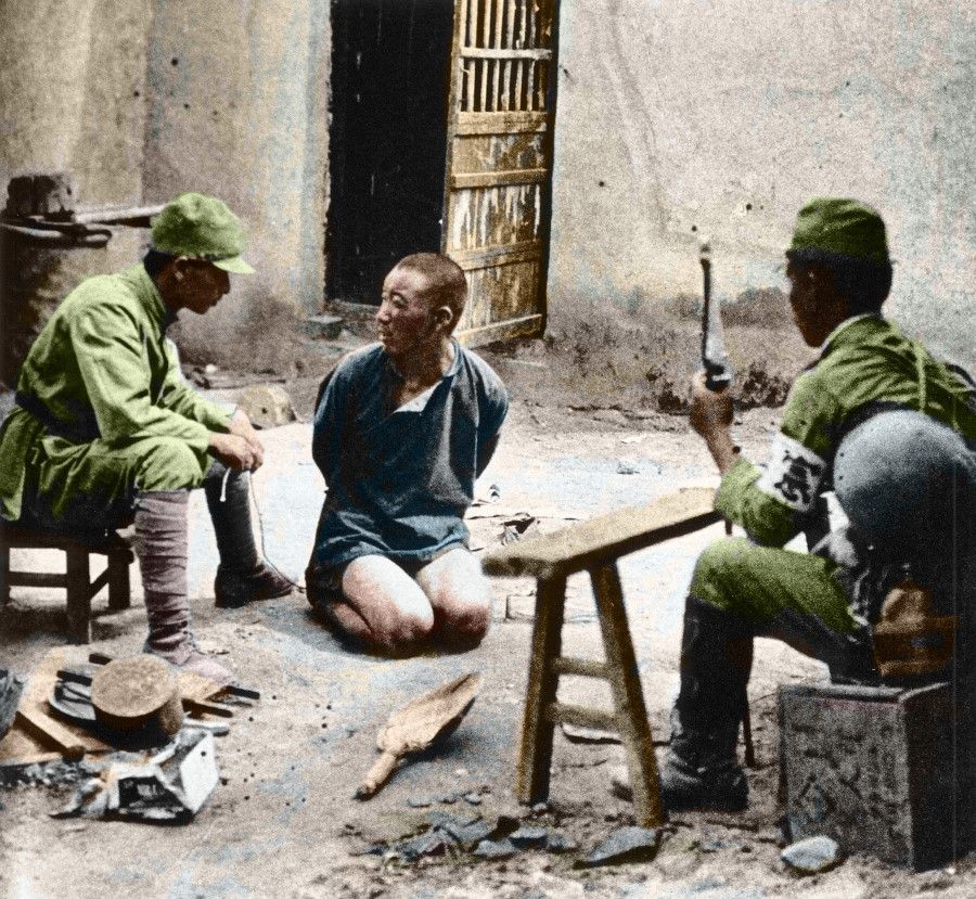 Japanese military police interrogating a Chinese soldier in a farmhouse near Tianjin, 8 August 1937. The Chinese man kneels on the ground with his hands bound behind him looking fearful and helpless, while a Japanese military officer with a samurai sword interrogates him. On the left is the Japanese interpreter. This photo was taken by the Japanese and was only released after the war.