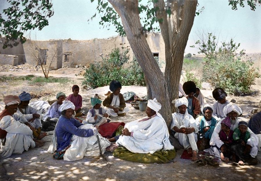 An all-male group chats under the shade of a tree, Tajikistan, early 20th century. Their clothing style is not identical; the men in the foreground are wearing loose white robes, with long cloths wrapped around their heads, while those behind the tree are wearing black sheepskin hats, and might be from a different tribe.