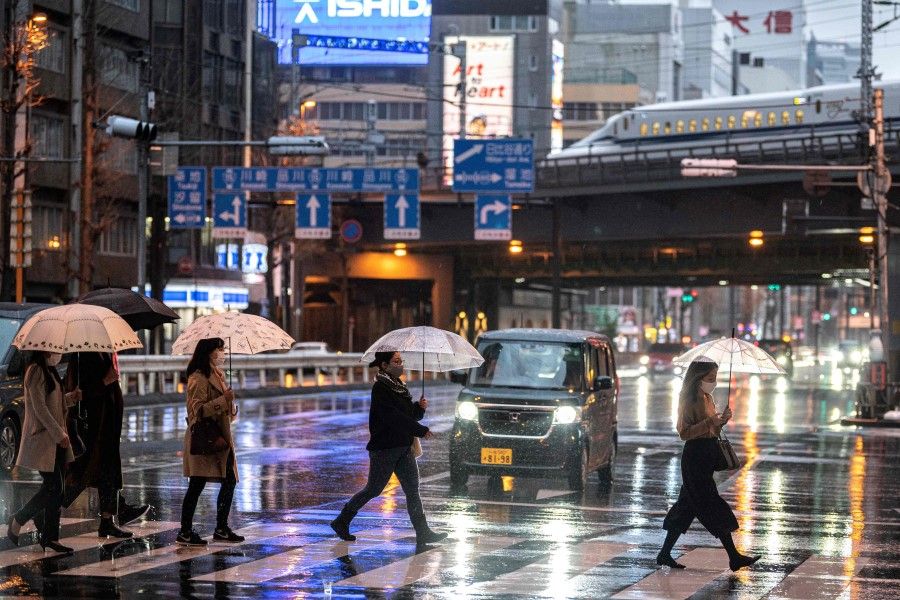 People cross a street under the rain at dusk while a shinkansen N700A series, or high speed bullet train, leaves Tokyo on 21 March 2021. (Charly Triballeau/AFP)