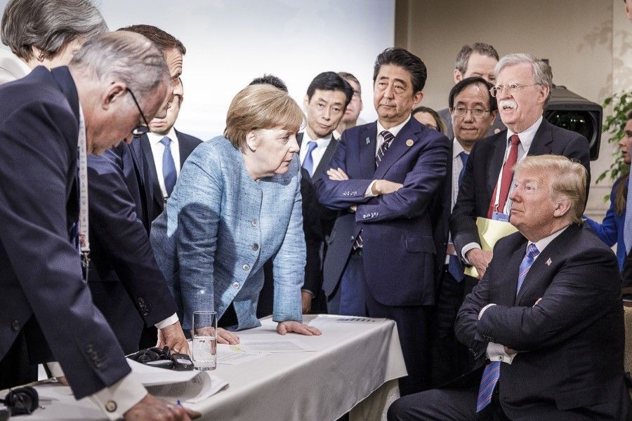 In this photo provided by the German Government Press Office (BPA), then Japanese Prime Minister Shinzo Abe (standing, in striped tie) looks on as German Chancellor Angela Merkel deliberates with US President Donald Trump on the sidelines of the official agenda on the second day of the G7 summit on 9 June 2018 in Charlevoix, Canada. (Jesco Denzel /Bundesregierung)