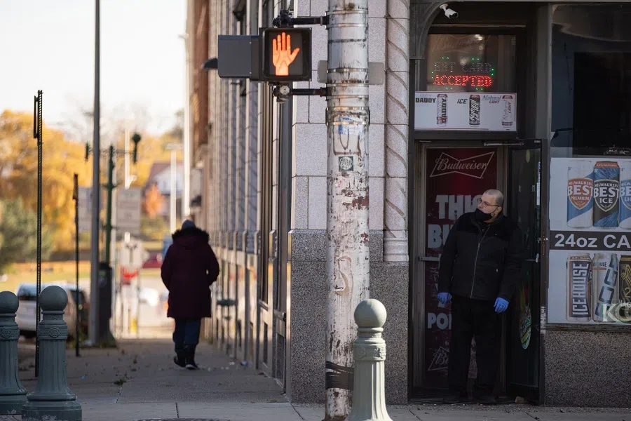 A pedestrian walks down the street as a store owner wearing a protective mask and gloves looks out the front door of a shop in downtown Pontiac, Michigan, US, on 31 October 2020. (Emily Elconin/Bloomberg)