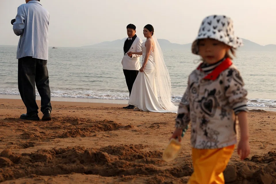A child plays with sand near a couple taking part in a pre-wedding photoshoot on a beach in Qingdao, Shandong province, China, on 21 April 2024. (Florence Lo/Reuters)