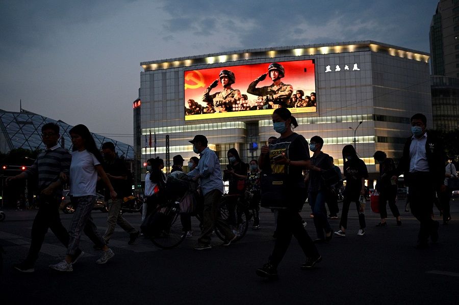 This photo taken on 18 May 2021 shows people walking along a street as military propaganda is displayed on a giant screen in Beijing, China. (Noel Celis/AFP)
