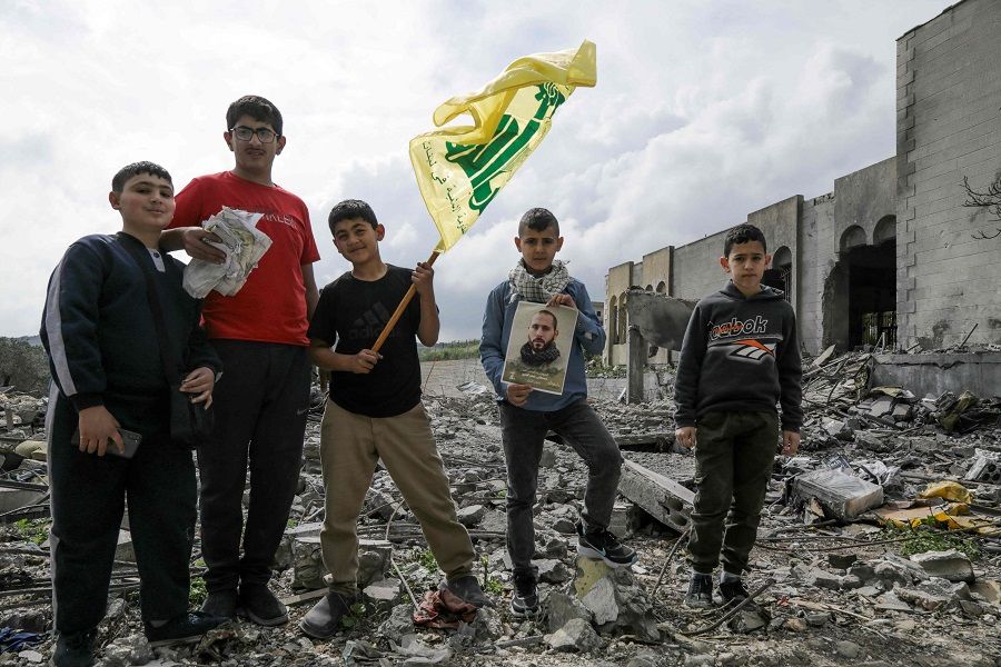 Boys pose with a Hezbollah flag above debris caused by previous Israeli fire as residents of the southern Lebanese village of Aita al-Shaab visit their deserted village on the occasion of a funeral of a Hezbollah fighter, on 9 April 2024, in southern Lebanon near the border with Israel. (Hasan Fneich/AFP)