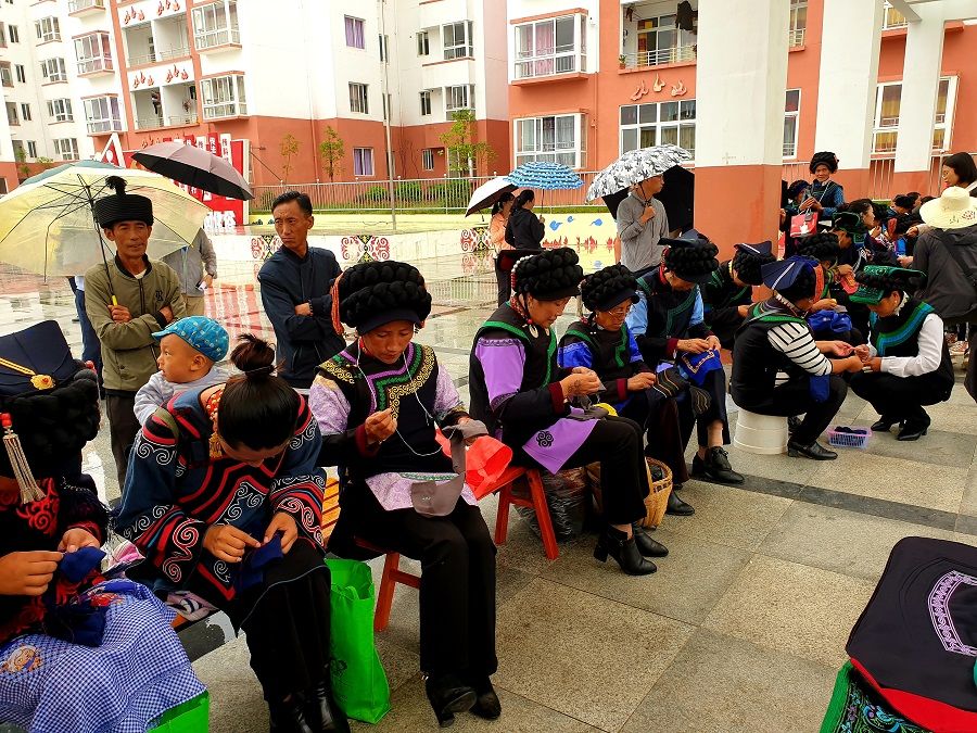 Yi women dressed in their traditional costumes are seen busying their hands with embroidery at the communal square of the Chengbei Thanksgiving Community. The government-built flats they have relocated to are seen in the background.