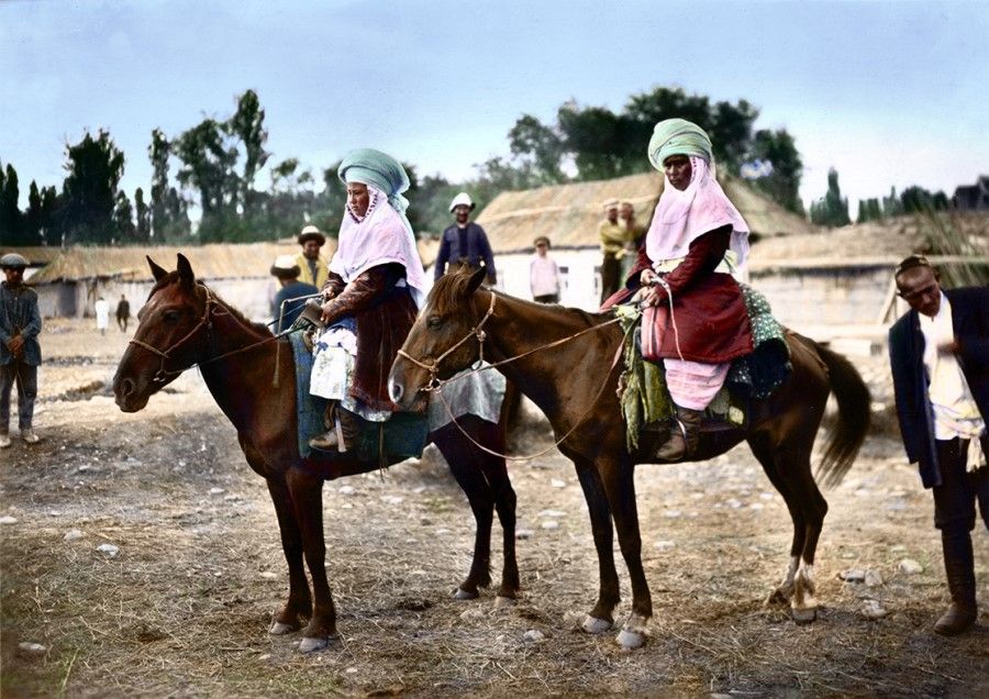 Tajikistan, early 20th century. Two women sit on horses, wearing long robes that cover their entire body, with light-coloured yet vibrant headscarves. Homes are built with thatched roofs, with walls built out of stone and mud.