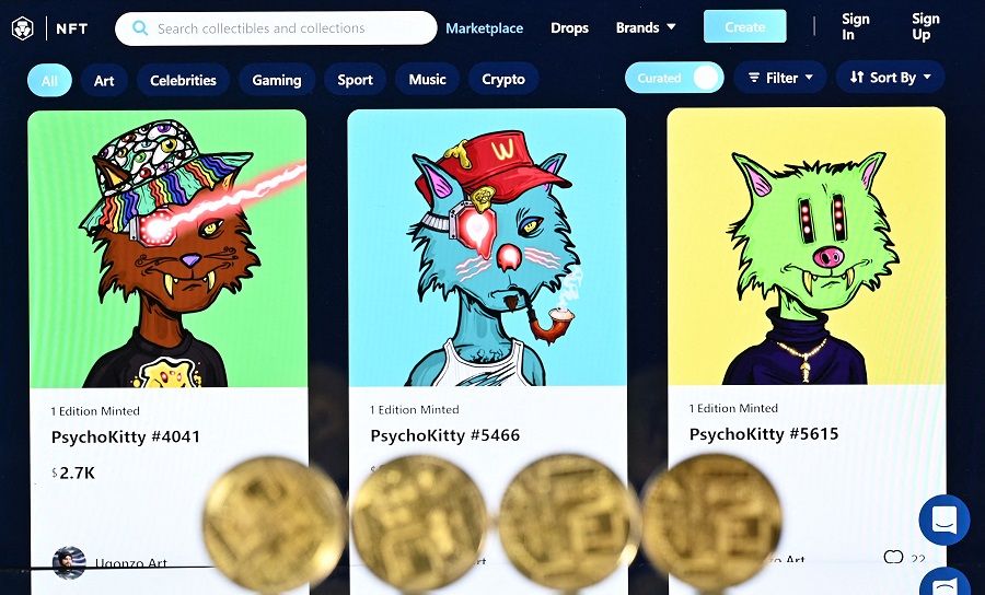 An illustration picture taken in London on 30 December 2021, shows gold plated souvenir cryptocurrency coins arranged beside a screen displaying a Crypto.com NFT (Non-Fungible Token) marketplace showing three PsychoKitty NFTs created by psychedelic artist Ugonzo. (Justin Tallis/AFP)