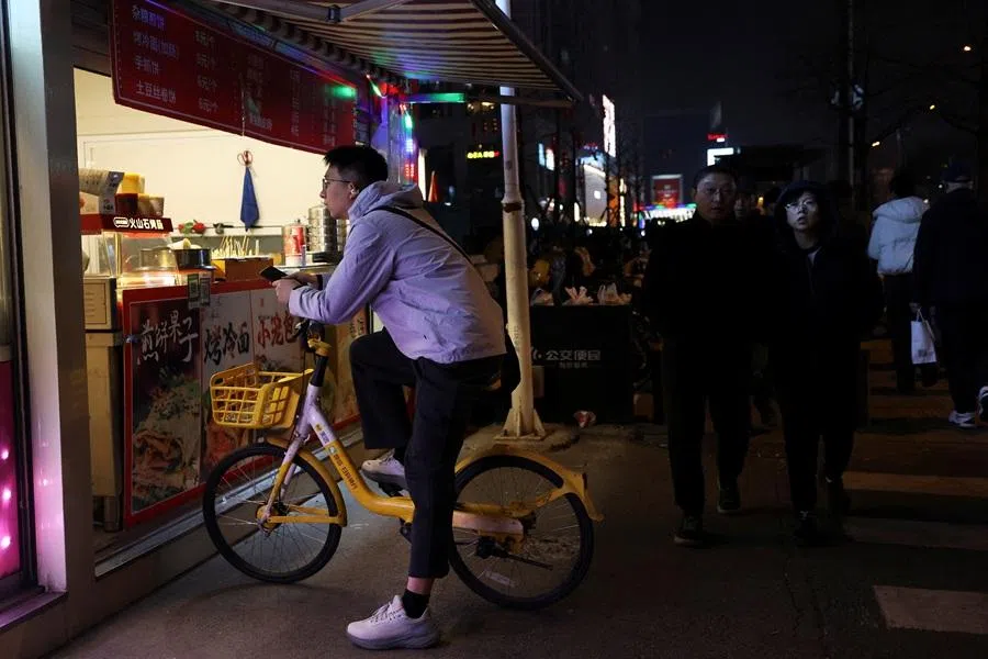 A man sitting on a shared bicycle waits for his order in front of a roadside food stall in Beijing, China, on 13 March 2026. (Florence Lo/Reuters)