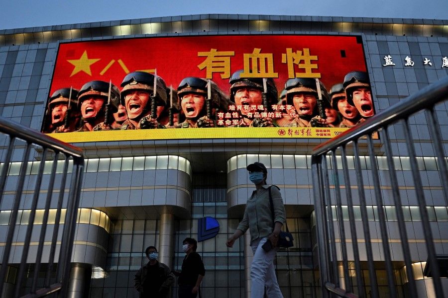 This photo taken on 18 May 2021, shows people walking along a street as military propaganda which reads "Courageous - Raise a new generation of spirited, capable, courageous and morally upright revolutionary soldiers" on a giant screen in Beijing. (Noel Celis/AFP)
