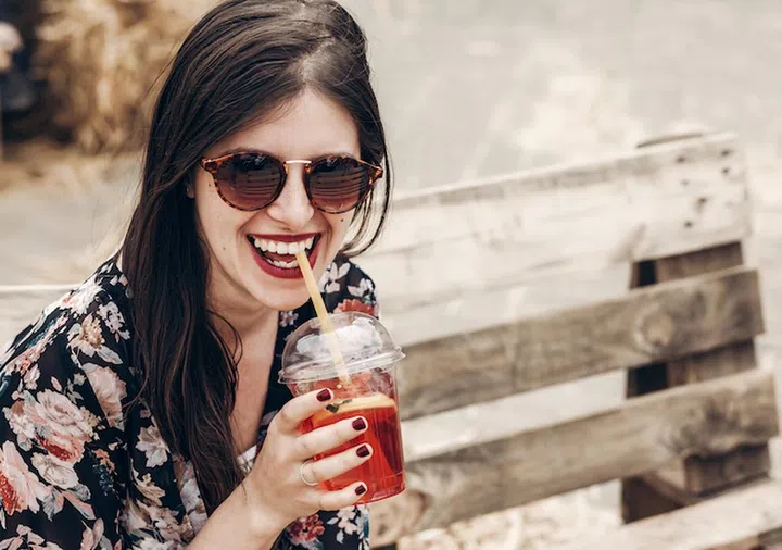 stylish hipster woman in sunglasses with red lips drinking lemonade. boho girl holding cocktail and smiling at street food festival. summertime. summer vacation travel. space for text