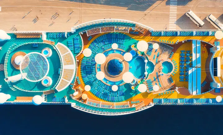 Aerial view of luxury cruise liner. Top view of swimming pool, people on sunbeds, umbrellas on the cruise ship in summer. View from above of relaxing people on deck chairs, blue water in pool, wooden deck. Resort