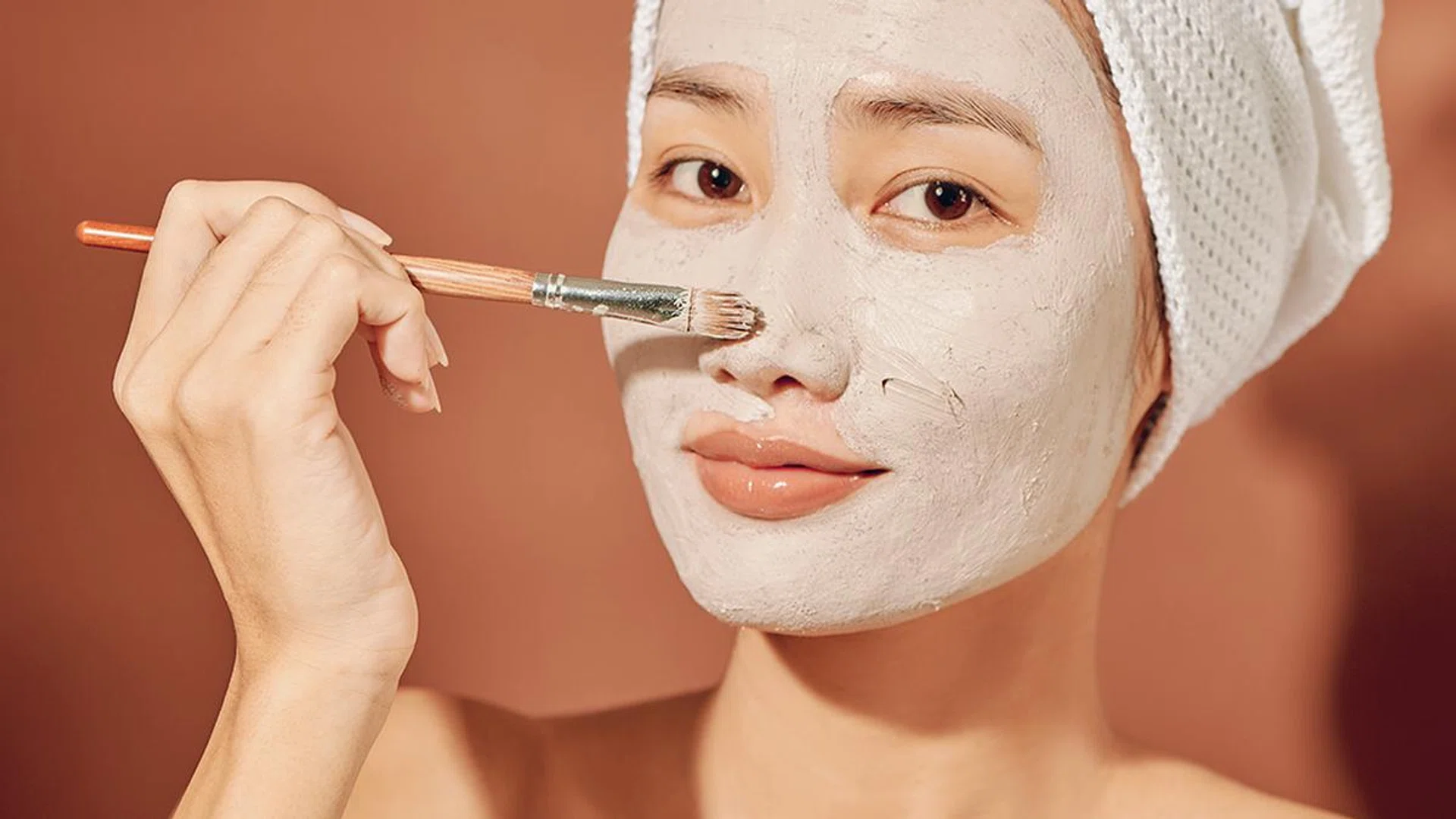 happy woman with a towel on her head apply a cleansing mask on her face
