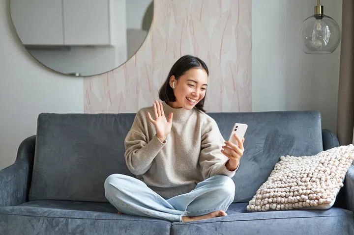 Portrait of asian woman sitting on sofa with smartphone, waving at mobile phone screen and waving at camera, video chat, talking to someone.