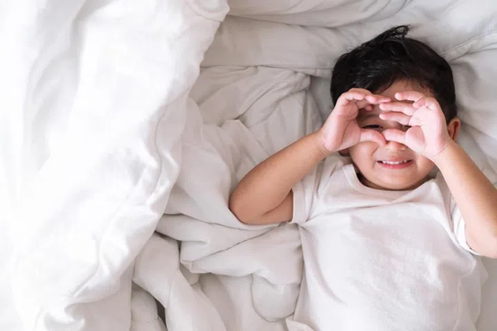 3 years old little cute Asian boy in white shirt at home on the bed, kid lying playing and smiling on white bed with pillow and blanket, top view with copy space for add text.