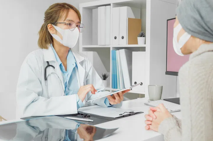Young Asian woman doctors wear protective mask medical results during consultation on tablet computer with female patient sick with cancer in health clinic. Medical health care concept.