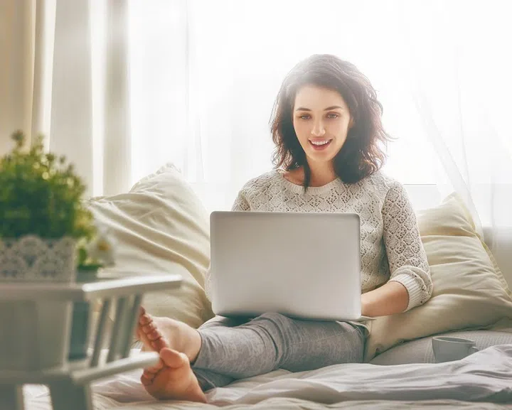 Happy casual beautiful woman working on a laptop sitting on the bed in the house.