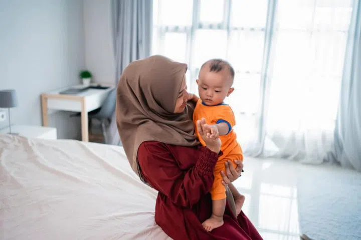 Asian moslem mother holding her little boy on lap when playing together on the bed