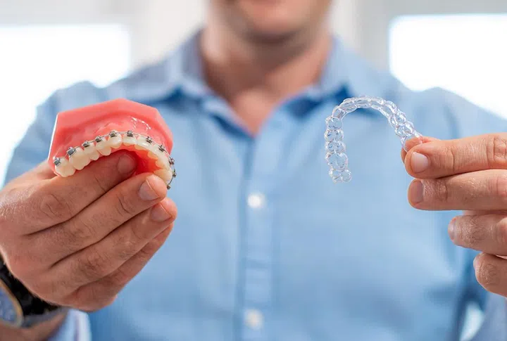 Dental care.Smiling orthodontist doctor holding aligners and braces in hand shows the difference between them
