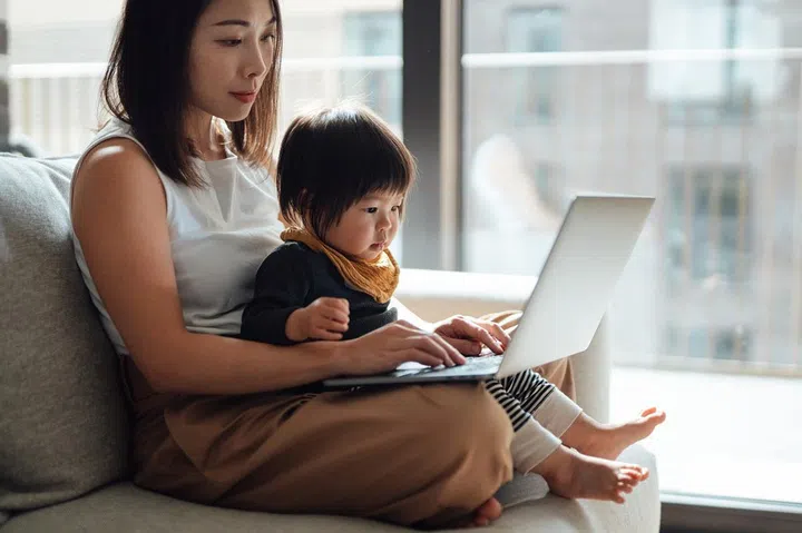 Young Asian mother working with laptop on the sofa while her little daughter sitting on her lap. Working mother. Parents working from home with kids. Adult online learning.