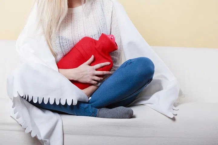 Painful periods and menstrual cramp problems concept. Woman having stomach cramps sitting on cofa feeling very unwell holding hot water bottle to feel some relief