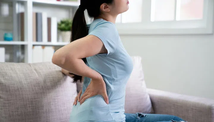 side view of unrecognized asian chinese woman in t shirt hands touching back suffer backache sitting on sofa at home. mother resting on couch in living room in day time while waist painful.