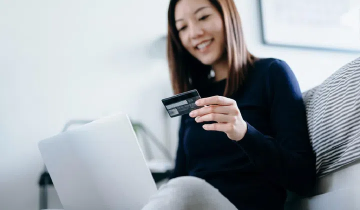 Beautiful smiling young Asian woman sitting on the floor in the bedroom, shopping online with laptop and making mobile payment with credit card on hand at cozy home. Technology makes life so much easier
