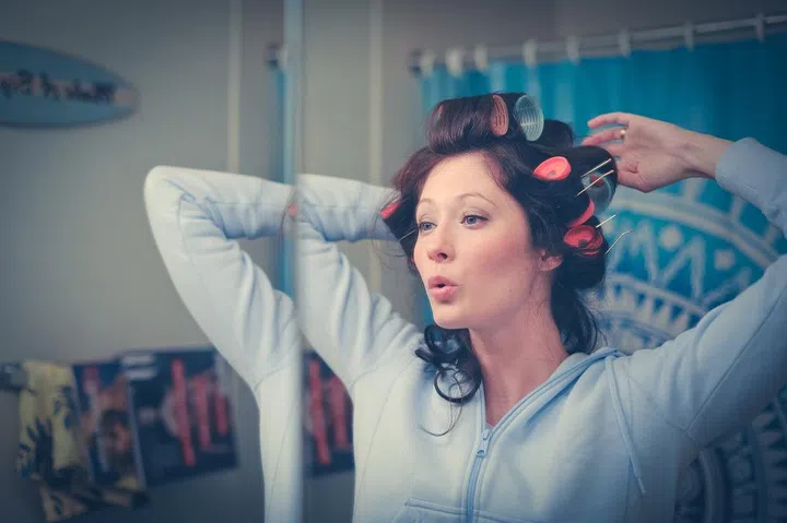 Young woman styling her hair with curlers, in blue hooded sweatshirt, singing or whistling in bathroom.