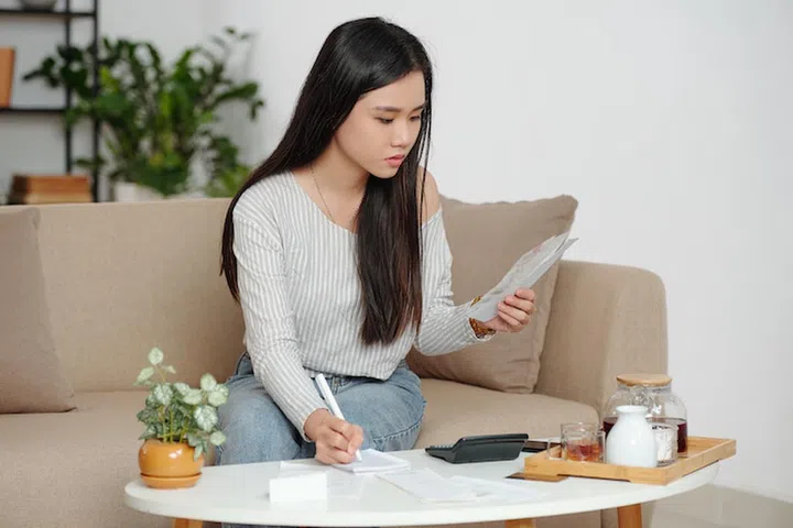Serious young Vietnamese woman calculating bank loan or rent payments and checking taxes when sitting on couch at home