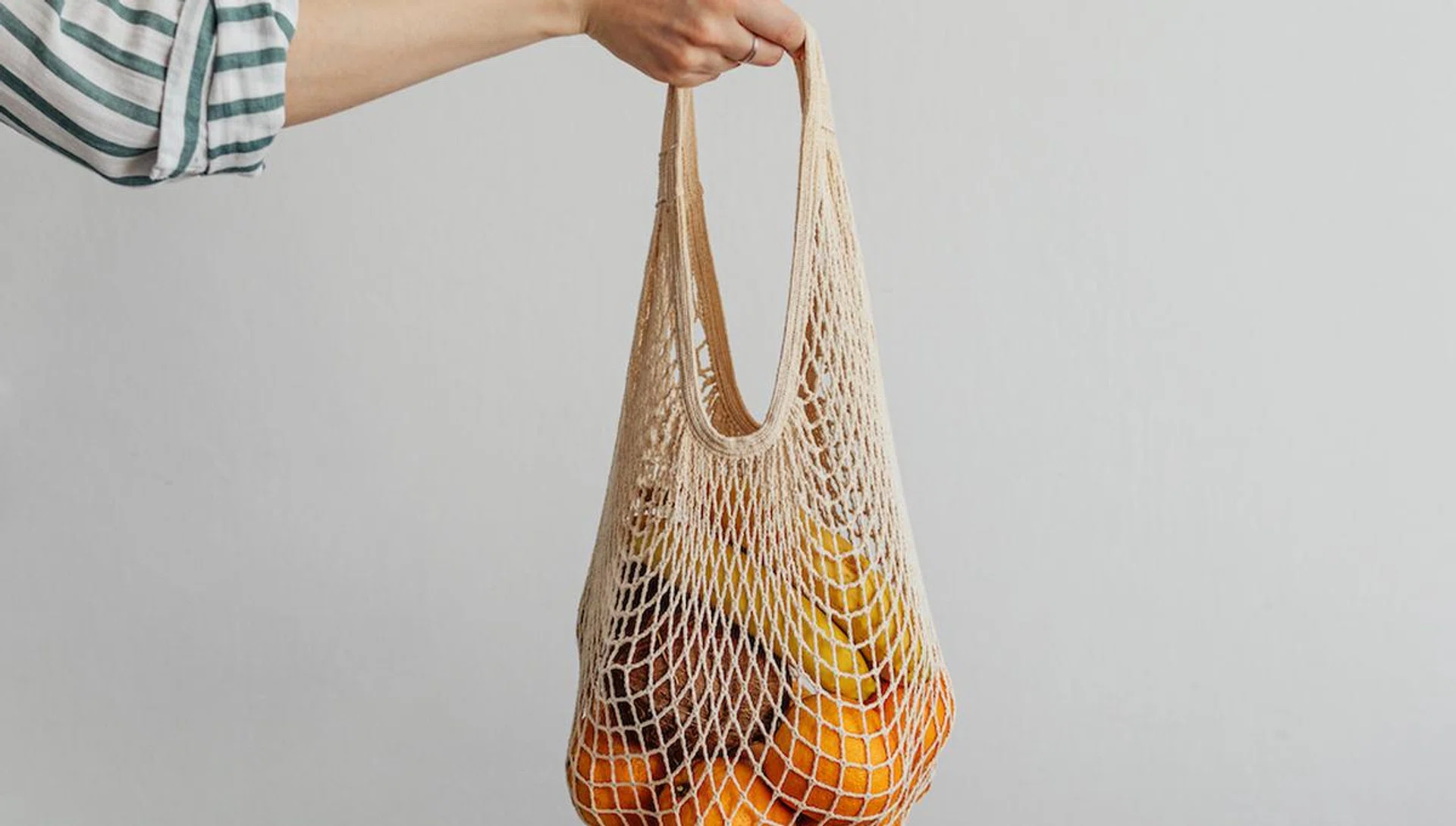 Mesh bag with fruits in female hand. Stylish young woman hand hold mesh shopping bag on light gray wall. Modern reusable shopping concept.