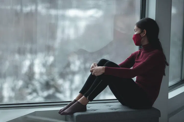 Winter depression because of coronavirus confinement. Sad Asian woman alone during city lockdown wearing face mask indoors at home covid. Anxiety, stress, mental health issues.