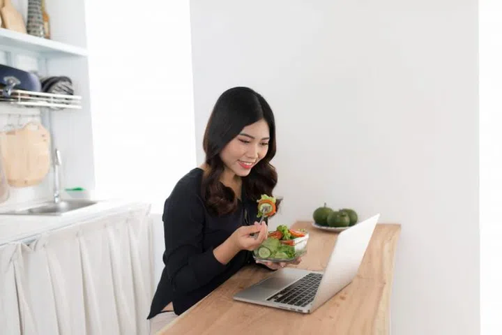 Young woman with salad working on laptop in the kitchen
