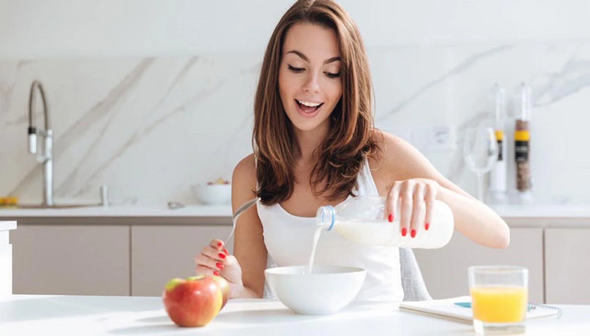 Happy joyful woman pouring milk into a bowl while sitting and having breakfast at the kitchen table