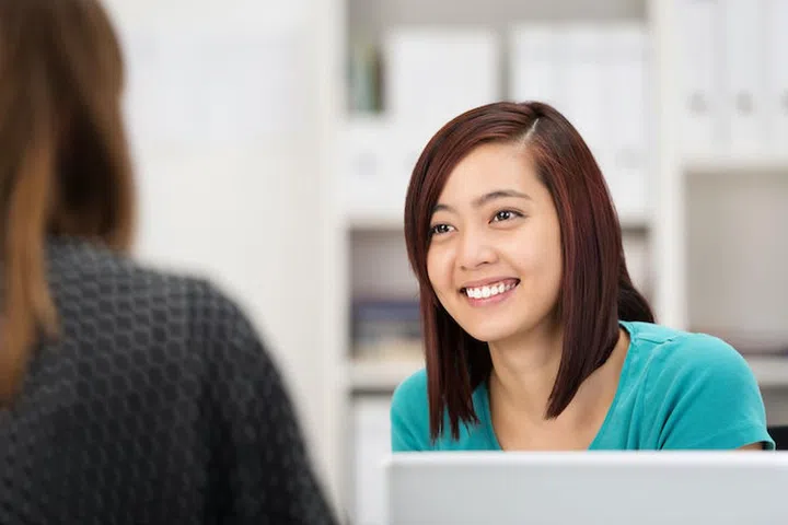 Smiling Asian businesswoman chatting to a female colleague as she sits at her desk in the office