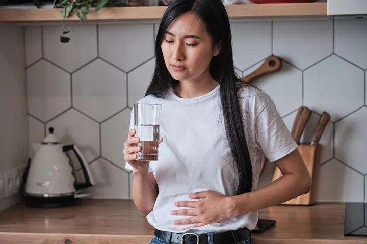 Sick young Asian woman standing in kitchen holding glass of water suffering from stomach ache, unhealthy girl feeling unwell during period, touching belly stomach having menstrual cramps