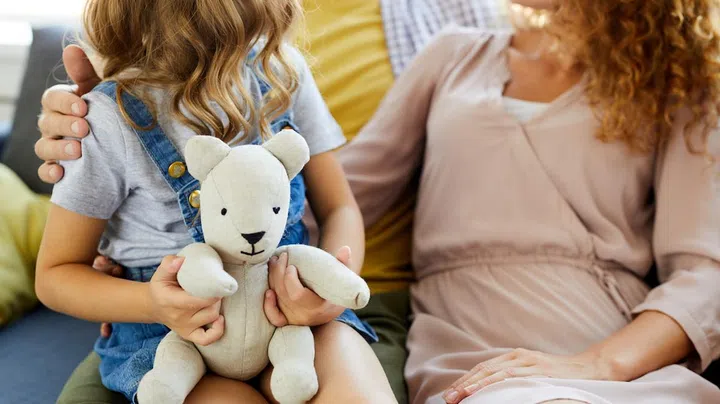 Little girl holding white teddybear with her mom and dad sitting near by
