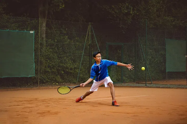 Asian boy performing a forehand during a tennis match on a clay court